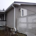 A gray building with vinyl siding, a white door, and white lattice fencing sits under a cloudy sky. There is a covered porch to the left and a white gutter along the roof and down the side of the house. Kentucky Builders And Excavating