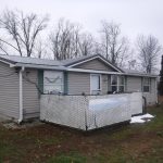 A single-story gray building with white trim, a small fenced porch, and a satellite dish on the roof, set in a yard with bare trees on a cloudy day. Kentucky Builders And Excavating