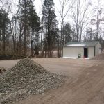 A gravel lot is under construction near a forested area. A large pile of rocks sits in the foreground, with excavating equipment, a metal shed, and dirt piles nearby. Leafless trees surround the scene. Kentucky Builders And Excavating