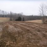 A muddy, plowed field stretches into the distance on a cloudy day, with a pole barn standing nearby, surrounded by leafless trees and patches of bare earth and grass in a rural landscape. Kentucky Builders And Excavating