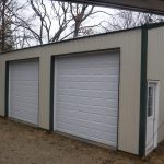 A beige metal pole barn building with three large white roll-up doors and a single white entry door on the right, surrounded by trees and dirt ground—perfect for storage or an excavating workspace. Kentucky Builders And Excavating