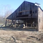 A large, weathered pole barn with missing boards and partial collapse stands on uneven ground, surrounded by debris from the old building, with bare trees and a clear sky in the background. Kentucky Builders And Excavating
