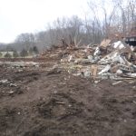 Debris from a collapsed pole barn lies scattered on muddy ground, with broken wood, concrete blocks, and metal roofing. Leafless trees and an open field are visible in the background under an overcast sky. Kentucky Builders And Excavating