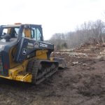 A John Deere 333G compact track loader stands on dirt at a demolition site, excavating piles of wood and debris from a collapsed building, with leafless trees visible beyond. Kentucky Builders And Excavating