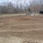 A wide dirt area with visible tire tracks from recent excavating, a pile of debris near the center, leafless trees in the background, and an open metal shed on the right. Kentucky Builders And Excavating