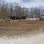A dirt lot with piles of debris in front of a weathered, open pole barn. Leafless trees surround the area, and a white fence runs along the right side, with a small excavating vehicle visible in the background. Kentucky Builders And Excavating
