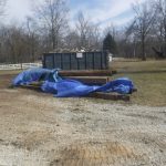 A dumpster filled with debris sits on a grassy area, surrounded by bare trees. Wooden beams and other materials for building a pole barn are covered with blue tarps in front. A white fence runs along the background. Kentucky Builders And Excavating