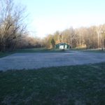 A gravel lot sits in the foreground of a grassy field with bare trees in the background. A small green pole barn building is visible near the tree line under a clear sky. Kentucky Builders And Excavating