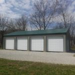 A large metal pole barn building with a green roof and four white roll-up doors sits on a gravel driveway, surrounded by grass and leafless trees under a partly cloudy sky. Kentucky Builders And Excavating