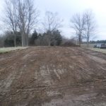 A freshly excavated and leveled patch of dirt sits in a grassy field, bordered by leafless trees under an overcast sky. A pickup truck and trailer are parked to the right, ready for building a pole barn. Kentucky Builders And Excavating