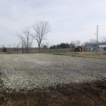 A large gravel pad is laid out on a rural property, ready for building or excavating, bordered by grass and bare trees, with farm buildings and a pole barn visible in the background under a cloudy sky. Kentucky Builders And Excavating