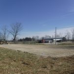 A construction site with a gravel road being built, a bulldozer on the left excavating, bare trees, and a few buildings—including a pole barn and a house—in the background under a clear blue sky. Kentucky Builders And Excavating
