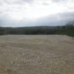 A blurry photo of an open, gravel-covered area where excavating is underway, with grass and trees in the background under a cloudy sky. Kentucky Builders And Excavating