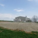 A beige, single-story house with a small shed and a nearby pole barn sits on a gravel lot surrounded by green grass and a few trees under a partly cloudy sky. Kentucky Builders And Excavating