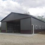 A large metal-sided pole barn with an open garage door and a white side door, set on a concrete foundation, sits next to a gravel area under a cloudy sky—ideal for building or excavating projects. Kentucky Builders And Excavating
