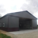 A large metal-sided pole barn with a wide open entrance, several small windows, and a gravel driveway, set in a rural area with grass and trees under a cloudy sky—ideal for building projects or excavating equipment storage. Kentucky Builders And Excavating