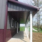 A covered porch with white pillars accents the side of a gray and maroon pole barn building, featuring a concrete walkway and a grassy area in the background. Kentucky Builders And Excavating