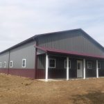 A large pole barn with gray metal walls, maroon trim, and a covered porch supported by white posts sits on bare earth, hinting at recent excavating work beneath a cloudy sky. Kentucky Builders And Excavating