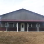 A large, brown metal pole barn with a maroon roof, white pillars, and a white door stands on a dirt lot, surrounded by grass and trees in the background. Kentucky Builders And Excavating