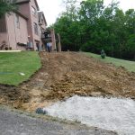 A muddy, sloped yard next to a brick house is under construction, with piles of dirt and excavating equipment on site. Two people stand at the top of the slope near the house, as trees line the background, possibly preparing for a building project. Kentucky Builders And Excavating