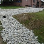 A sloped driveway under construction beside a brick house, with excavating equipment nearby, large white rocks and rebar frames in place, and building materials scattered across grass and dirt on either side. Kentucky Builders And Excavating