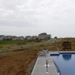A backyard pool with lounge chairs sits beside a muddy, unfinished yard where excavating work is underway. In the background, a suburban street curves past newly built houses and the frame of a pole barn on a cloudy day. Kentucky Builders And Excavating