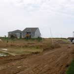 A muddy construction site with two gray houses in the background, a white pickup truck parked on a dirt road, and some excavating equipment and puddles scattered around the area under a cloudy sky. Kentucky Builders And Excavating