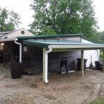 A person bends over under a green metal pole barn attached to a weathered wooden shed. The ground is wet and muddy, and there are trees and a grassy area in the background. Kentucky Builders And Excavating