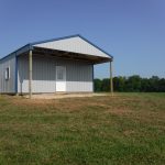 A small, light gray pole barn with a blue roof sits on a grassy field. It has a covered front porch supported by wooden posts and a white door facing forward, with trees and clear sky in the background. Kentucky Builders And Excavating
