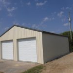 A beige metal pole barn with two white roll-up doors sits on a concrete driveway under a partly cloudy blue sky. There is a utility pole and some greenery nearby, making this building ideal for storage or workshops. Kentucky Builders And Excavating