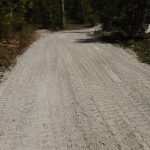 A gravel road curves through a wooded area, with trees and patches of grass lining both sides under daylight. Visible tire tracks mark the surface, and a pole barn building stands nearby, surrounded by greenery. Kentucky Builders And Excavating