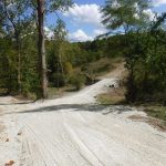 A gravel road winds downhill through a wooded area with green trees under a partly cloudy sky. Some rocks and a small corrugated metal roof near the road’s edge suggest recent excavating for a building project. Kentucky Builders And Excavating