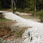 A gravel road crosses a shallow ditch in a wooded area, with a culvert pipe visible under the road and piles of rocks on the side—ideal conditions for excavating before constructing a building. Trees and dense vegetation surround the scene. Kentucky Builders And Excavating
