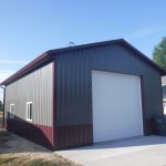 A metal pole barn or storage building with a white roll-up door, gray and maroon siding, two small windows on the side, and a trash bin by the entrance sits on a concrete driveway beside a lawn. Great for building or excavating projects. Kentucky Builders And Excavating