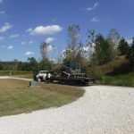 A black skid steer loader, used for excavating, is being transported on a flatbed trailer pulled by a white pickup truck along a gravel driveway near a grassy lawn, trees, and a pole barn under a blue sky with scattered clouds. Kentucky Builders And Excavating