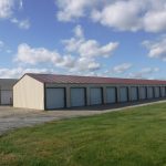 A row of storage units with white roll-up doors and a metal roof stands beside a gravel driveway under a partly cloudy sky, resembling a modern pole barn building, with green grass in the foreground. Kentucky Builders And Excavating
