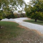A curving gravel driveway under construction winds through a grassy yard, bordered by trees with green and yellow leaves. Piles of white gravel are visible on the left side as part of the excavating work for a future pole barn. Kentucky Builders And Excavating