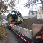 A small excavator is excavating dirt onto a flatbed trailer parked in a narrow alleyway, with traffic cones on the trailer and houses visible in the background. Kentucky Builders And Excavating