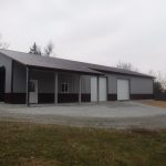 A large metal pole barn with a gray roof, three white garage doors, a small white door, and a window sits among gravel and sparse trees on an overcast day. Perfect for building projects or excavating equipment storage. Kentucky Builders And Excavating