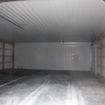 Interior of an empty, unfinished pole barn or storage building with a concrete floor, exposed wooden beams on the walls, and a small door in the back. Tire tracks from recent excavating work are visible across the floor. Kentucky Builders And Excavating