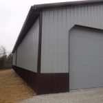 A large gray metal pole barn with maroon trim, featuring a closed overhead garage door. The building sits on a gravel path with dry grass and trees visible in the background under a cloudy sky. Kentucky Builders And Excavating