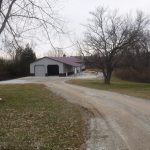 A gravel driveway curves toward a large pole barn with a red roof and white walls, surrounded by leafless trees and grassy areas on a cloudy day—an ideal spot for building or excavating projects. Kentucky Builders And Excavating