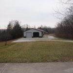 A small white metal pole barn with two large doors sits at the end of a gravel driveway, surrounded by grass and leafless trees on a cloudy day—perfect for storage, building projects, or even light excavating work. Kentucky Builders And Excavating