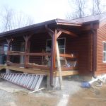A wooden cabin with a covered front porch is under construction, alongside a new pole barn. Construction materials, scaffolding, and tools are scattered around the site. Leafless trees provide a quiet backdrop to the ongoing building work. Kentucky Builders And Excavating
