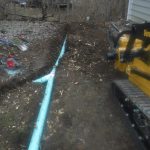 A trench next to a house with a large light blue pipe being installed; a yellow Vermeer excavating machine is on the right, and tools and dirt are scattered around the building site. Kentucky Builders And Excavating