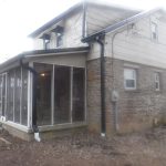 A two-story house with stone and siding exterior, screened porch, visible downspouts, and a yard with bare ground—ideal for future building or excavating projects. Another house and scattered leaves are visible in the background. Kentucky Builders And Excavating