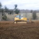 A yellow bulldozer is excavating soil on a muddy field, preparing the ground for building a pole barn, with trees in the background and a pine branch in the foreground on a cloudy day. Kentucky Builders And Excavating