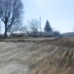 A cleared, gravel-covered construction site with tire tracks in the foreground, a building foundation and pole barn in the middle ground, a semi-trailer in the background, and bare trees on a sunny day. Kentucky Builders And Excavating