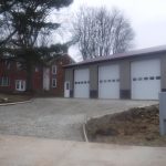 A large pole barn garage with three white doors sits beside a two-story brick house, surrounded by trees, gravel, and a newly constructed driveway on a cloudy day. Kentucky Builders And Excavating