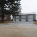 A modern pole barn with three large garage doors and a side entrance sits next to a large pine tree and a red-brick house on a paved driveway under an overcast sky. Kentucky Builders And Excavating